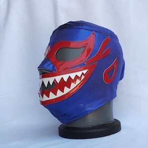 Peut inclure: Un masque de lucha libre bleu avec des détails rouges. Le masque présente une bouche et des yeux rouges brillants avec des dents blanches. Il est présenté sur une tête de mannequin noire sur fond blanc.