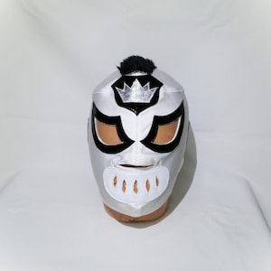 Puede incluir: Una máscara de lucha libre plateada y negra con un emblema de corona. La máscara presenta detalles negros alrededor de los ojos y una abertura para la boca. Una pequeña mata de pelo negro está en la parte superior de la máscara.