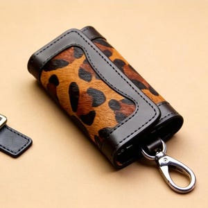 Può includere: Un portachiavi con un motivo leopardato. Il portachiavi è realizzato in pelle marrone e nera con una chiusura color argento. È incluso anche un cinturino separato in pelle nera con fibbia argentata.