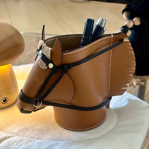Könnte beinhalten: Ein brauner Schreibtisch-Organizer aus Leder in Form eines Pferdekopfes mit schwarzen Zaumzeugdetails und einem geflochtenen Akzent. Der Organizer enthält Stifte und Bleistifte. Der Pferdekopf ist etwa 15 cm hoch.