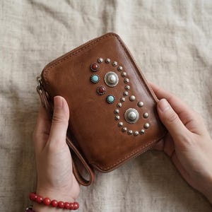 May include: Brown leather wallet with a zip closure, adorned with silver studs and turquoise and red accents. The wallet has a wrist strap and is held by two hands.