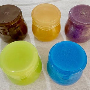 May include: Five small, round, translucent jars with matching lids, arranged in a group. The jars are colored dark brown, yellow, purple with glitter, lime green, and bright blue. Each container has a smooth, rounded body and a flat, circular lid.