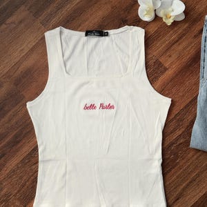May include: A white ribbed vest top with a square neckline featuring the text "bette porter" embroidered in red thread across the centre chest. The garment includes a black interior neck label with white text and a size indicator.
