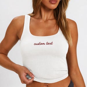 May include: A white ribbed vest top featuring a scoop neckline and wide shoulder straps. The centre of the chest displays the words custom text embroidered in a dark red cursive font. The garment has a fitted silhouette and a straight hemline.