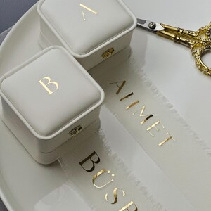 Op de afbeelding: Twee witte vierkante ringdozen met gouden letters, een met de letter "A" en de andere met "B". Een wit lint met de namen "AHMET" en "BÜSRA" in gouden letters is ook aanwezig. Een gouden schaar ligt op een wit bord.