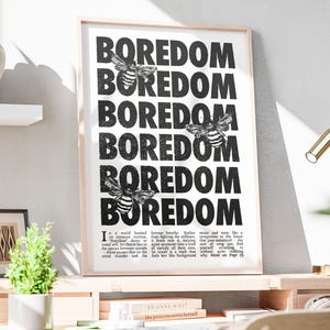 Puede incluir: Un cuadro enmarcado con la palabra "BOREDOM" repetida en negrita, fuente negra, intercalada con ilustraciones de abejas. La impresión también incluye un párrafo de texto en la parte inferior. La obra de arte se exhibe en un escritorio de madera.
