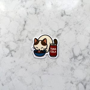 May include: A sticker featuring a cartoon cat with brown markings, eating from a blue bowl. Beside the cat is a red can labeled "TUNA COLA". The cat has its tongue out, and the sticker has a white background.