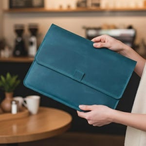Puede incluir: Funda para portátil de cuero color turquesa con cierre de solapa y una pestaña rectangular. La funda la sostiene una persona en un café. El fondo incluye una mesa de madera, una cafetera y una planta en un jarrón.