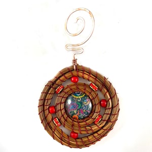 May include: A handcrafted ornament featuring a circular woven design with a central paisley-patterned glass cabochon. The ornament is adorned with red beads and crystals, and a copper wire spiral for hanging.
