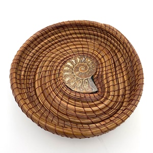 May include: A handcrafted, circular basket woven from natural pine needles, featuring a spiral design. A fossilized ammonite is embedded in the center. The basket is brown with a reddish-brown accent.