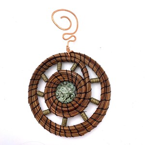 May include: A circular ornament crafted from coiled brown pine needles bound with black thread, featuring a central green and white paisley patterned cabochon, metallic gold-toned spacer beads, and a copper wire spiral hanger at the top.