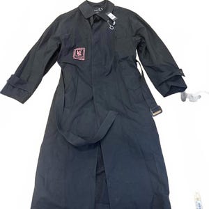 Peut inclure: Longue veste noire avec un col, des manches longues et une ceinture. La veste présente un écusson rose et noir sur la poitrine et une étiquette attachée au col. La veste est posée sur une surface blanche.