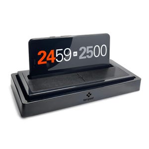 May include: A black rectangular charging dock with a glossy finish and the word tempest printed on the front. A smartphone is positioned horizontally in the dock, displaying the numbers 2459 and 2500 in orange, white, and grey on its screen.