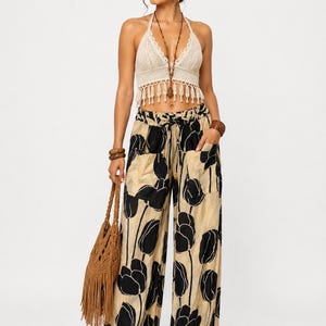May include: A beige crochet halter top with fringe trim paired with wide-leg trousers featuring a black tulip print on a beige background. The trousers have a drawstring waist and two large front pockets. A brown woven bag with fringe is included.