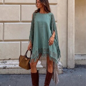 May include: A seafoam green poncho-style dress with fringe trim and patterned accents. The dress is paired with brown leather knee-high boots and a brown handbag. The outfit is suitable for casual wear.