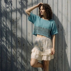 May include: A teal crop top with a geometric design and matching shorts. The shorts have a floral pattern and a ruffled lace hem. The model is standing barefoot in front of a weathered metal wall.