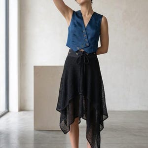 May include: A sleeveless blue satin vest with a V-neckline and a row of buttons, paired with a black high-waisted skirt featuring a textured, semi-sheer fabric, an asymmetrical handkerchief hemline, and a drawstring waist with metal eyelets.