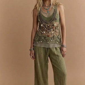 May include: Olive green tank top with intricate bead and shell embellishments, paired with matching wide-leg pants. The top features a mesh overlay and floral accents. A layered necklace and bracelets complete the bohemian style.