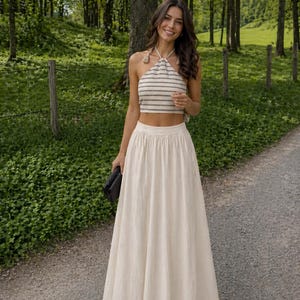 May include: A woman wearing a cream-colored maxi skirt and a halter top with black and white stripes. The outfit is paired with a black clutch. The background is a forest with a path.