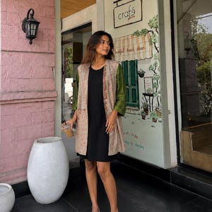 May include: A woman wearing a black dress and a long, open-front jacket with tan and green patterned sleeves. She is standing outside a building with a mural that says "crafts galerie". She is wearing beige flats and holding a beverage.