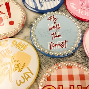 May include: A collection of round, decorative buttons with various designs and text. Some buttons feature pearl bead edging. Colors include red, blue, yellow, and pink. Text includes "Kappa Kappa Gamma", "Fly with the finest", and "CHIC!"