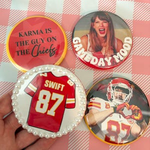 May include: Four round buttons with red and white designs. One button says "Karma is the guy on the Chiefs". Another says "Gameday Mood" with a photo. A third button has a red jersey with "SWIFT 87". The last button has a football player.