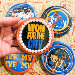 May include: A collection of round buttons featuring basketball imagery. One button has the text "WON FOR THE CITY" with pearl embellishments. Other buttons display team photos and player action shots. The buttons are in shades of blue, orange, and yellow.