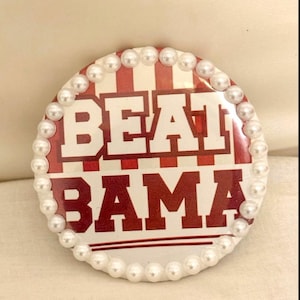 May include: A round button with the words "BEAT BAMA" in white block letters on a red and white striped background. The button is edged with small, white faux pearls.