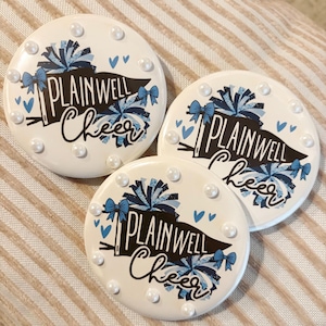 May include: Three circular white buttons decorated with small white pearls along the edges. Each button features a black pennant flag with the text "PLAINWELL" and the word "Cheer" in script, surrounded by blue pom-poms, bows, and heart icons.