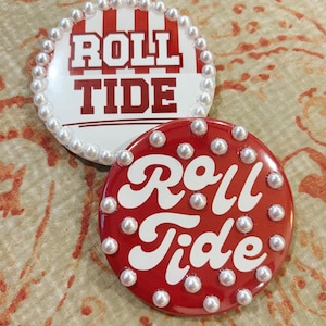 May include: Two circular pin-back buttons. The top button features red and white vertical stripes with the text "ROLL TIDE" in red, bordered by white faux pearls. The bottom button is solid red with white script text "Roll Tide" and white faux pearls.