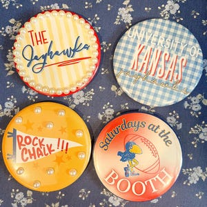 May include: Four round buttons with text and designs. One button has "THE Jayhawks" in red and blue, with a pearl border. Another says "University of Kansas Jayhawks" on a blue gingham background. A third says "ROCK CHALK !!!" and the last says "Saturdays at the BOOTH".
