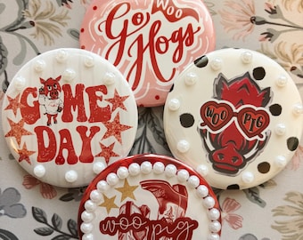 May include: Four circular pinback buttons featuring sports-themed designs. One is pink with red text reading Go Woo Hogs. Another is white with red stars, a hog mascot, and Game Day text. One features a hog in heart glasses, and one has cowboy boots.