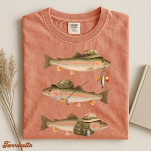 Terracotta Comfort Colors® shirt flat lay showing three rainbow trout with detailed fishing gear like camo hats, vest, and tackle.