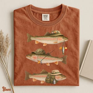 Yam Comfort Colors® flat lay shirt featuring three trout with fishing gear, camo hats, lures, and a vest. Ideal for outdoorsy gifts.