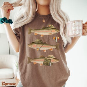 Woman wearing espresso Comfort Colors® shirt with three rainbow trout illustrated in full fisherman costume with rod and vest.