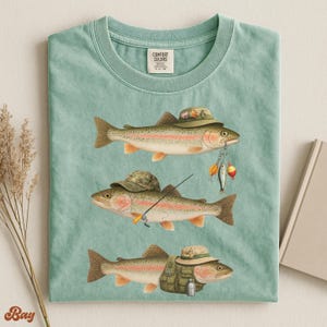 Soft green Comfort Colors® shirt laid flat, displaying three trout with detailed fisherman gear including rod, bait, hat, and vest.