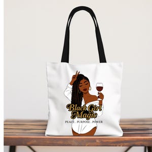 Puede incluir: Bolso tote blanco con asas negras. Presenta un gr&aacute;fico de una mujer negra sosteniendo una copa de vino y haciendo el signo de la paz. El texto "Black Girl Magic" est&aacute; en oro, con "PEACE. PURPOSE. POWER" debajo.