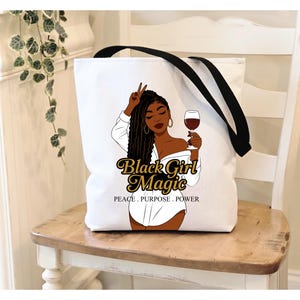 Puede incluir: Bolso tote blanco con correas negras. Presenta un gr&aacute;fico de una mujer negra sosteniendo una copa de vino. El texto "Black Girl Magic" est&aacute; en oro, con "PEACE. PURPOSE. POWER" debajo.