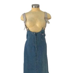 May include: A denim overall dress with a long skirt and side slit. The dress has adjustable striped straps and a front pocket. The dress is displayed on a mannequin.