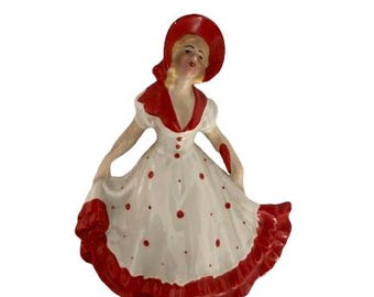 May include: A ceramic figurine of a woman in a white dress with red polka dots and trim. She wears a matching red hat and has blonde hair. The figurine is in a dancing pose.