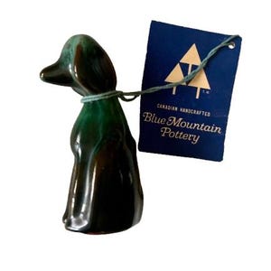 May include: A ceramic dog figurine with a glossy, dark green and brown finish. The figurine is attached to a blue tag with the text "Blue Mountain Pottery" and a logo of three trees. The figurine is approximately 4 inches tall.