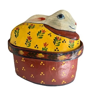 Puede incluir: Una caja de papel maché pintada a mano con forma de conejo. El conejo es blanco con un ojo azul y está acostado en una cesta con estampado floral amarillo y rojo. La cesta es roja con puntos amarillos.