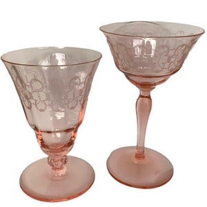May include: Two pink glass goblets with a delicate etched floral design. The goblets have a stemmed base and a wide bowl.