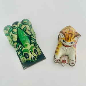Pode incluir: Dois brinquedos de lata vintage, um sapo verde e um gato laranja e branco. O sapo está deitado de costas com as patas no ar. O gato está sentado com as patas enfiadas debaixo do corpo.