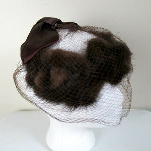 Puede incluir: Un sombrero de piel marrón con velo de red marrón y un lazo de satén marrón.