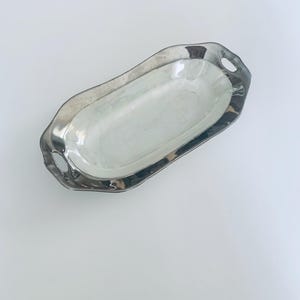 以下が含まれることがあります： 白い内側と2つの取っ手を持つ銀製のサービングディッシュ。ディッシュは楕円形で、波打った縁があります。