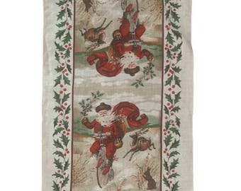 May include: A rectangular Christmas-themed fabric with a repeating illustration of Santa Claus riding a bicycle. The design features a border of holly and berries. The Santa illustrations are in red and white, set against a winter landscape.