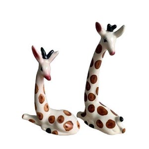 May include: Two ceramic giraffe figurines. The giraffes have white bodies with brown spots, black accents, and pink details on the ears and nose. One giraffe is lying down, and the other is sitting up.