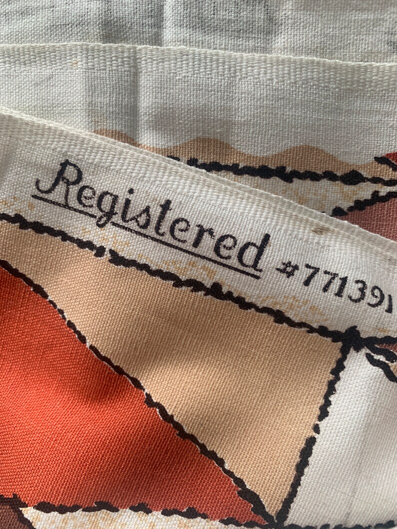 Peut inclure: Gros plan sur un tissu blanc avec un motif g&eacute;om&eacute;trique brun et orange. Le tissu a une bordure noire et le texte "Registered #771391" est imprim&eacute; sur le tissu.