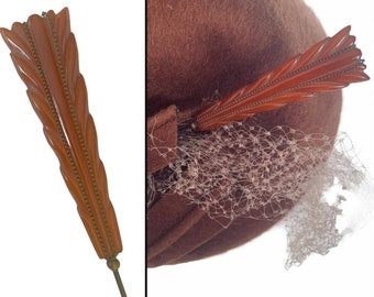 Puede incluir: Un sombrero marrón con un broche decorativo en forma de pluma. El broche es de color naranja cálido con un diseño detallado y un ribete color latón. Una delicada velo de red está adjunto al sombrero.